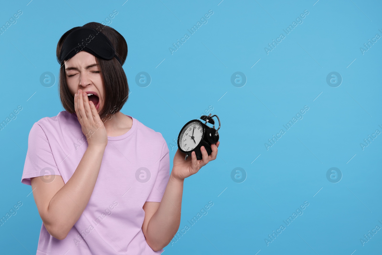 Photo of Sleepy young woman with sleep mask and alarm clock yawning on light blue background, space for text. Insomnia problem