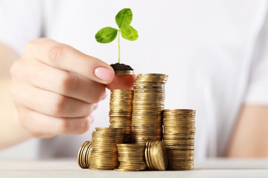 Woman putting coin with green sprout onto stack at white table, closeup. Investment concept