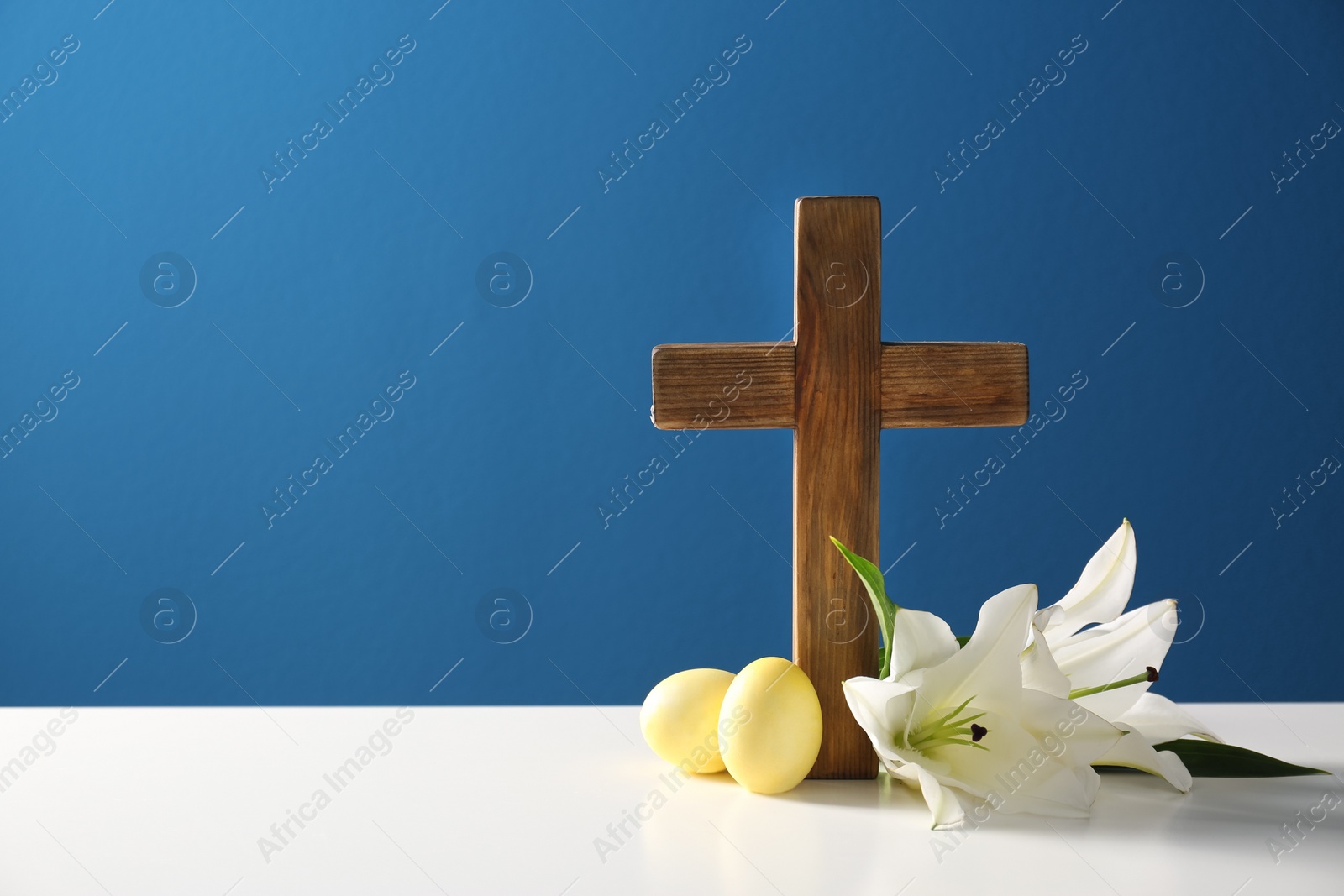 Photo of Wooden cross, Easter eggs and blossom lilies on table against color background, space for text