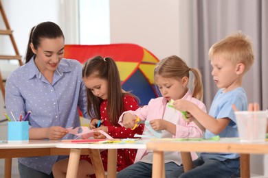 Photo of Nursery teacher and cute little children making toys from color paper at desks in kindergarten. Playtime activities
