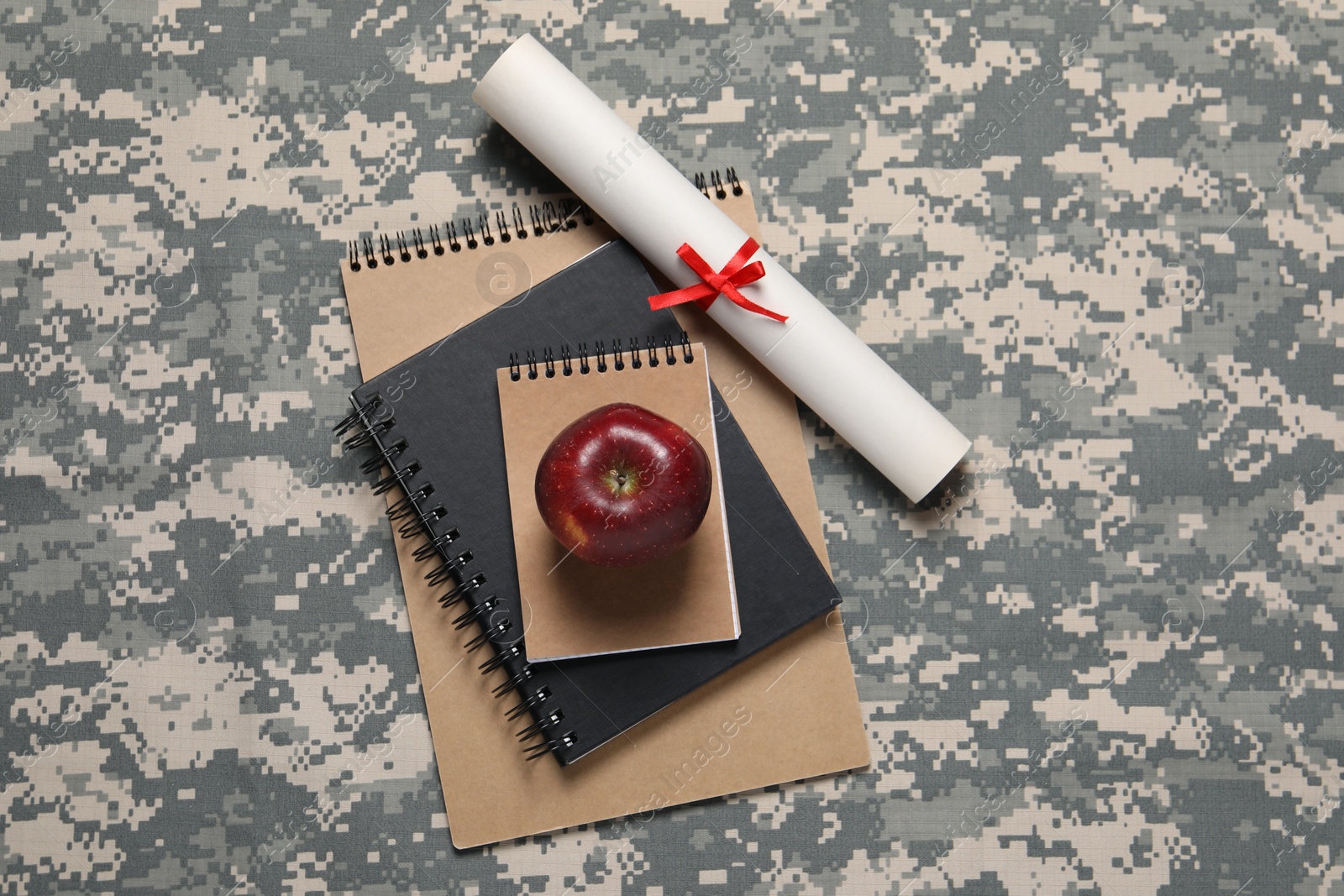 Photo of Notebooks, apple and diploma on camouflage background, flat lay. Military education