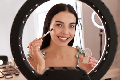 Photo of Beautiful young woman applying highlighter with brush indoors, view through ring lamp