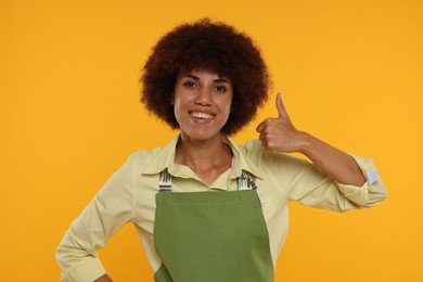 Photo of Happy young woman in apron showing thumb up on orange background