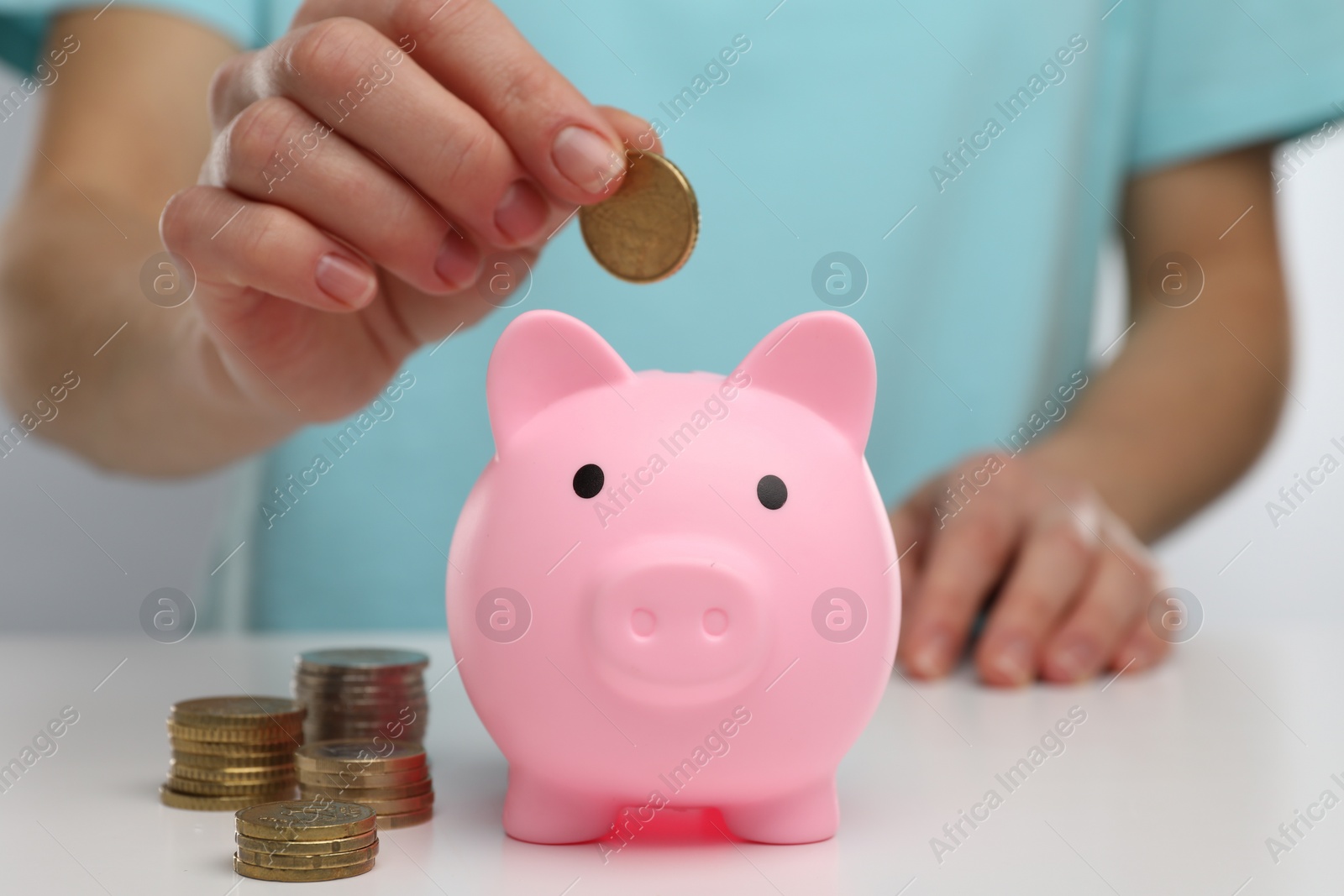 Photo of Woman putting coin into pink piggy bank at white table, closeup