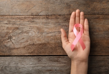 Woman holding pink ribbon on wooden background, top view with space for text. Breast cancer awareness concept