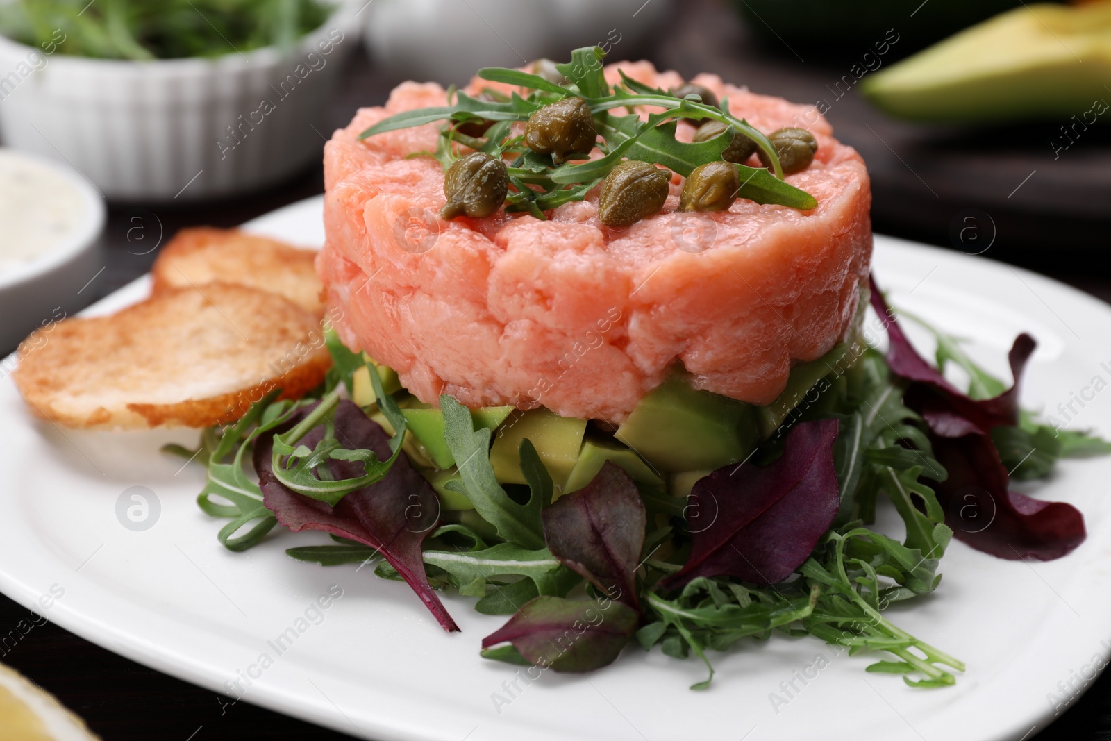 Photo of Tasty salmon tartare with avocado, greens and croutons on table, closeup