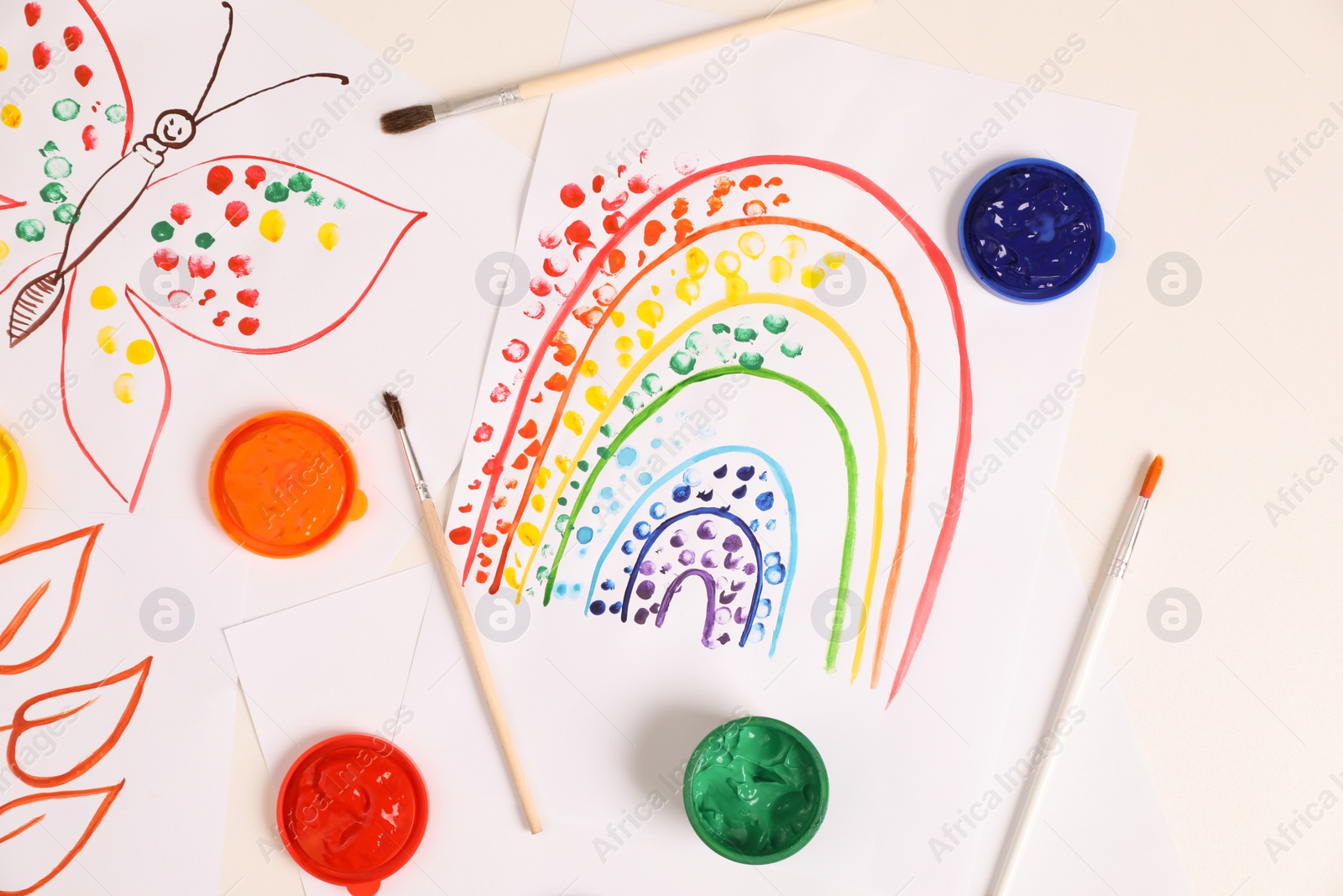 Photo of Cute children's drawings, brushes and set of paints on white table, flat lay