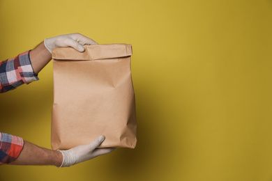 Photo of Courier in protective gloves holding paper bag on yellow background, closeup with space for text. Food delivery service during coronavirus quarantine