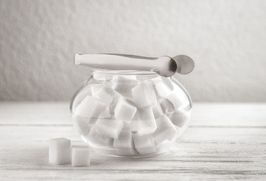 Photo of Refined sugar cubes in glass bowl and tongs on white wooden table