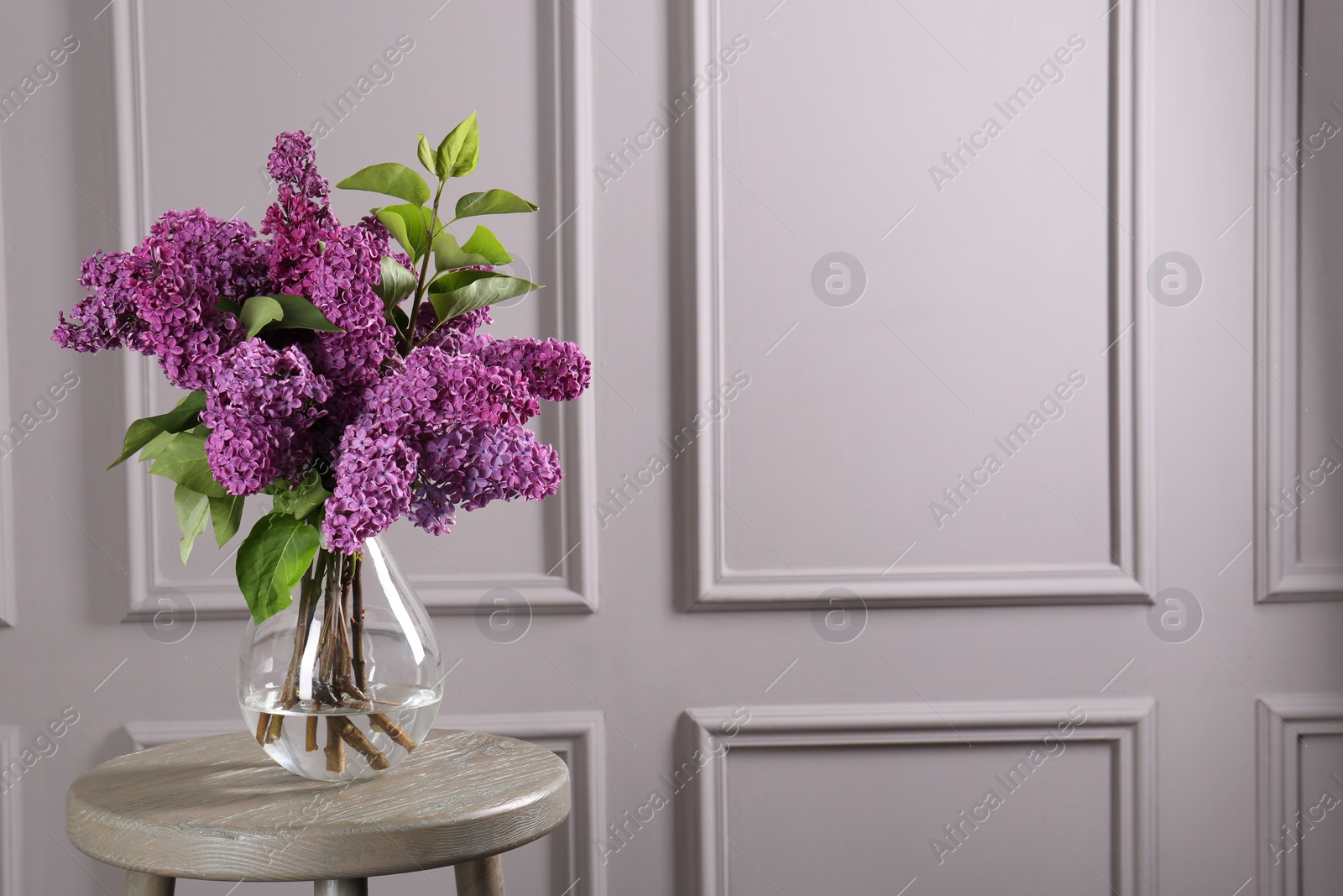 Photo of Beautiful lilac flowers in vase on wooden table near light wall, space for text