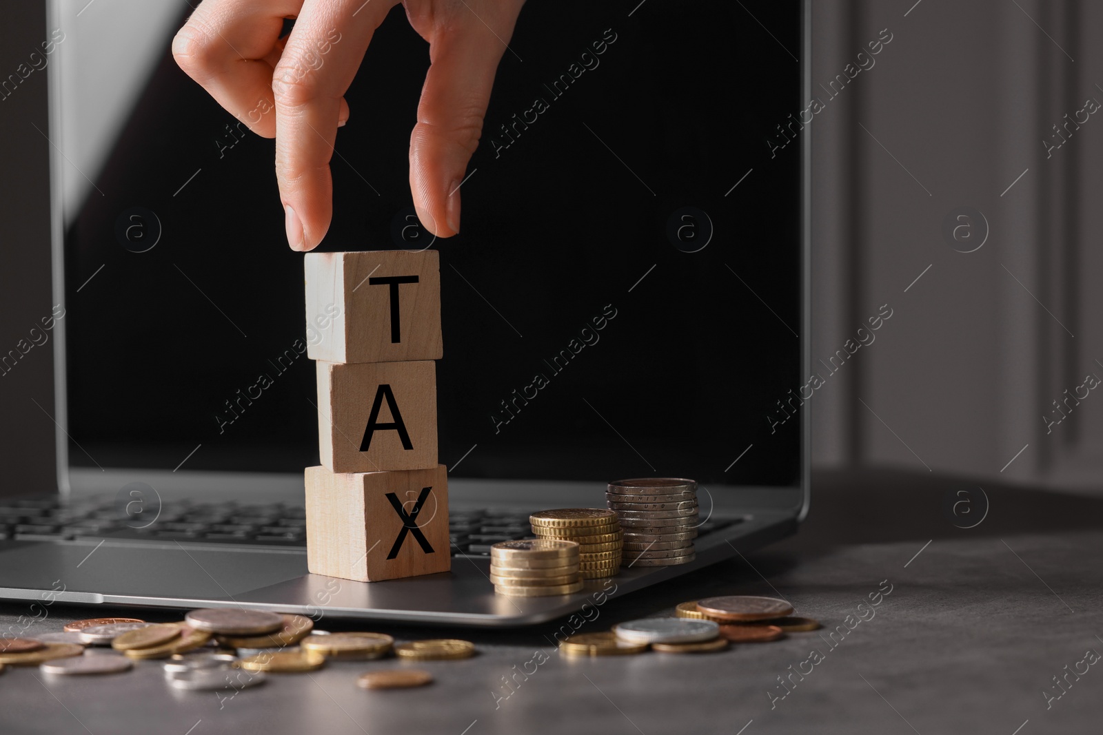 Photo of Woman with word Tax made of wooden cubes, laptop and coins at grey table, closeup. Space for text