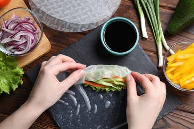 Photo of Woman wrapping spring roll at wooden table with products, top view