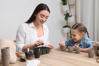 Mother and her daughter planting vegetable seeds into peat pots with soil at wooden table indoors