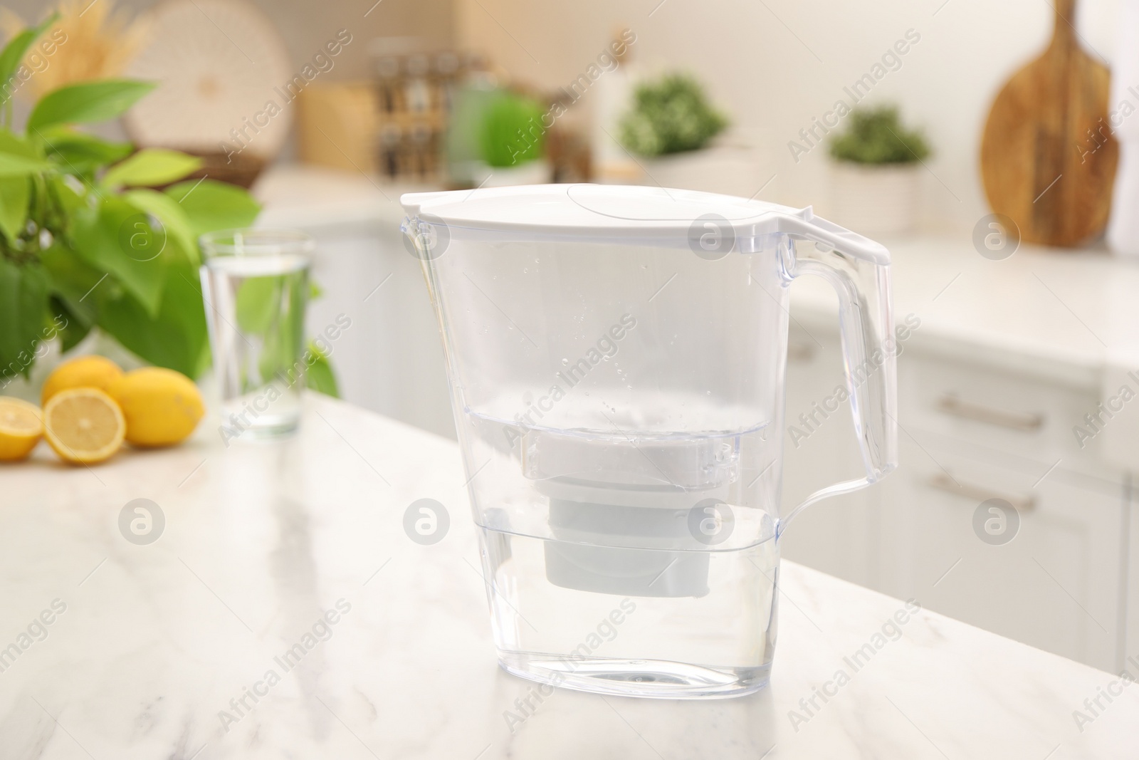 Photo of Water filter jug, glass and lemons on white marble table in kitchen