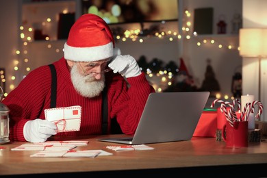Photo of Santa Claus using laptop and holding letter at his workplace in room decorated for Christmas