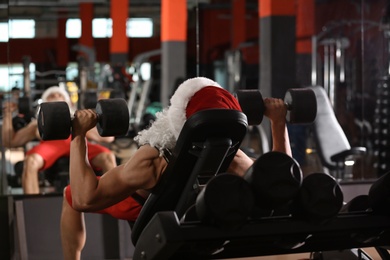 Young muscular man in Santa costume training with dumbbells at modern gym