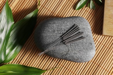 Photo of Stone with acupuncture needles and green leaves on bamboo mat, flat lay