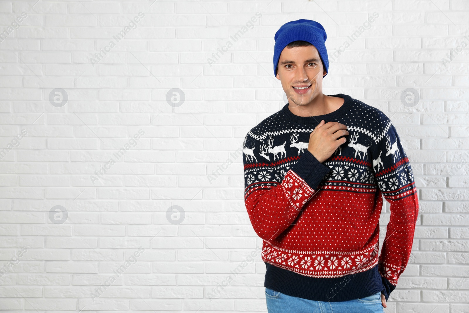 Photo of Portrait of young man in Christmas sweater and hat near white brick wall. Space for text