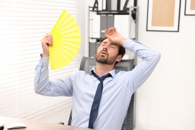 Bearded businessman waving yellow hand fan to cool himself at table in office