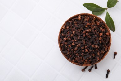 Aromatic cloves in bowl and green leaves on white tiled table, flat lay. Space for text