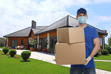 Courier in protective mask and gloves with cardboard boxes near house. Delivery service during coronavirus quarantine