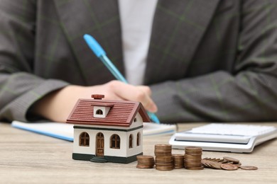 Photo of Woman writing at wooden table, focus on house model and stacked coins