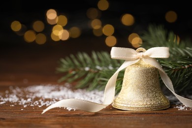 Photo of Bell with bow, fir branches and artificial snow on wooden table against blurred background, closeup. Christmas decor
