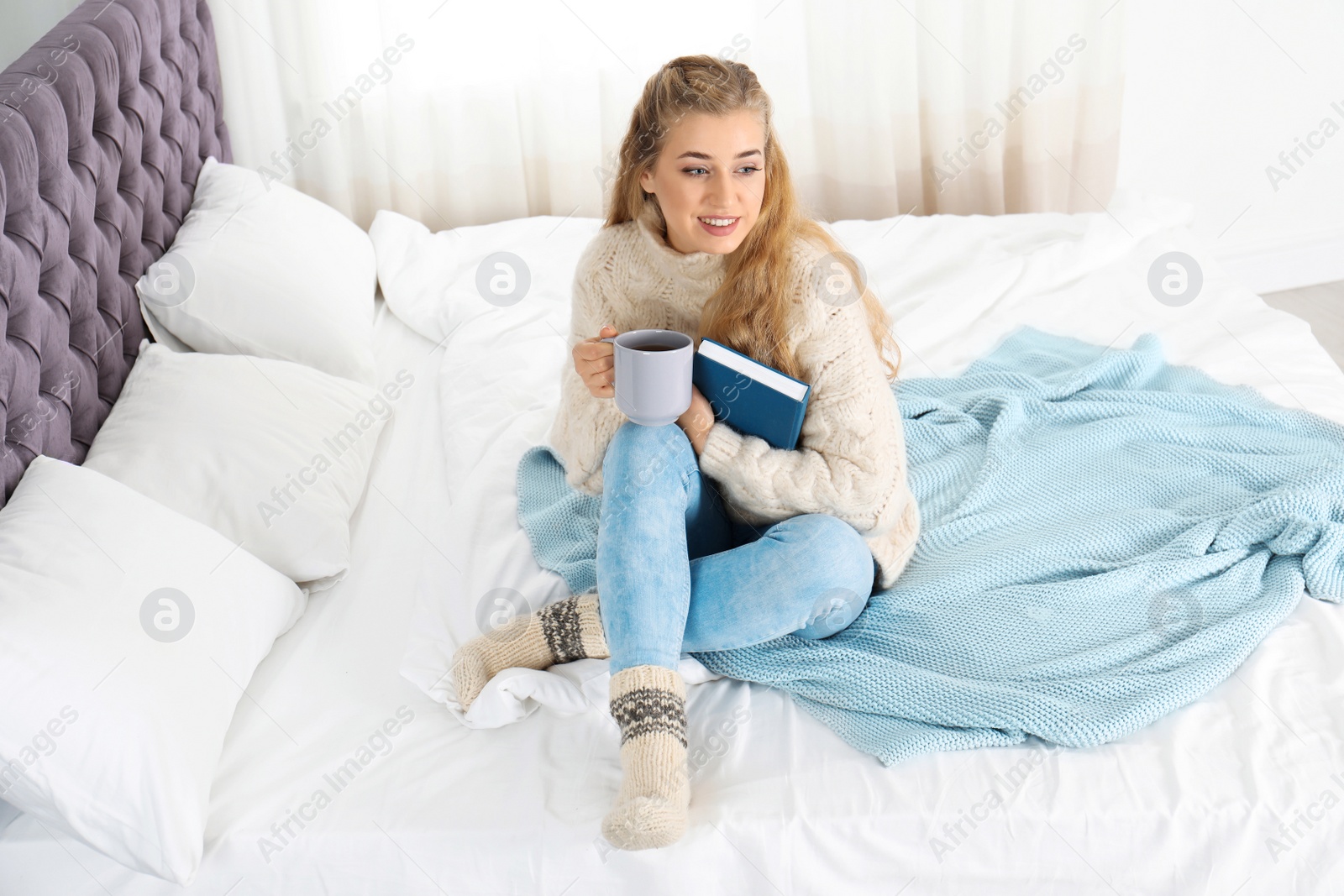Photo of Attractive young woman in cozy warm sweater with cup of hot drink and book sitting on bed at home