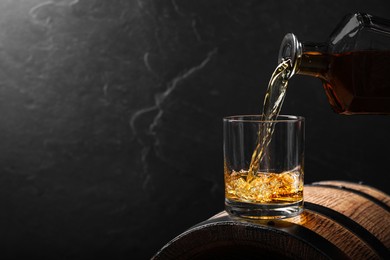 Photo of Pouring whiskey into glass from bottle on wooden barrel against grey background, space for text