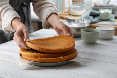 Photo of Woman stacking homemade sponge cakes at white wooden table in kitchen, closeup