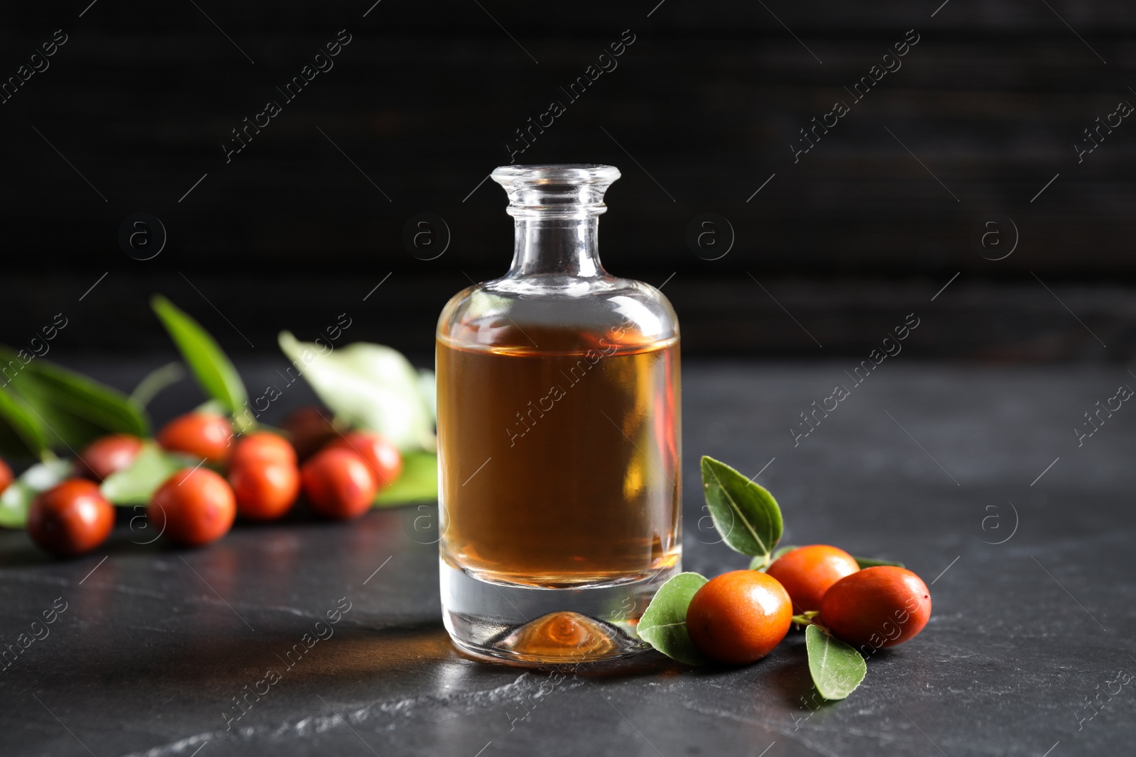 Photo of Glass bottle with jojoba oil and seeds on grey stone table against dark background