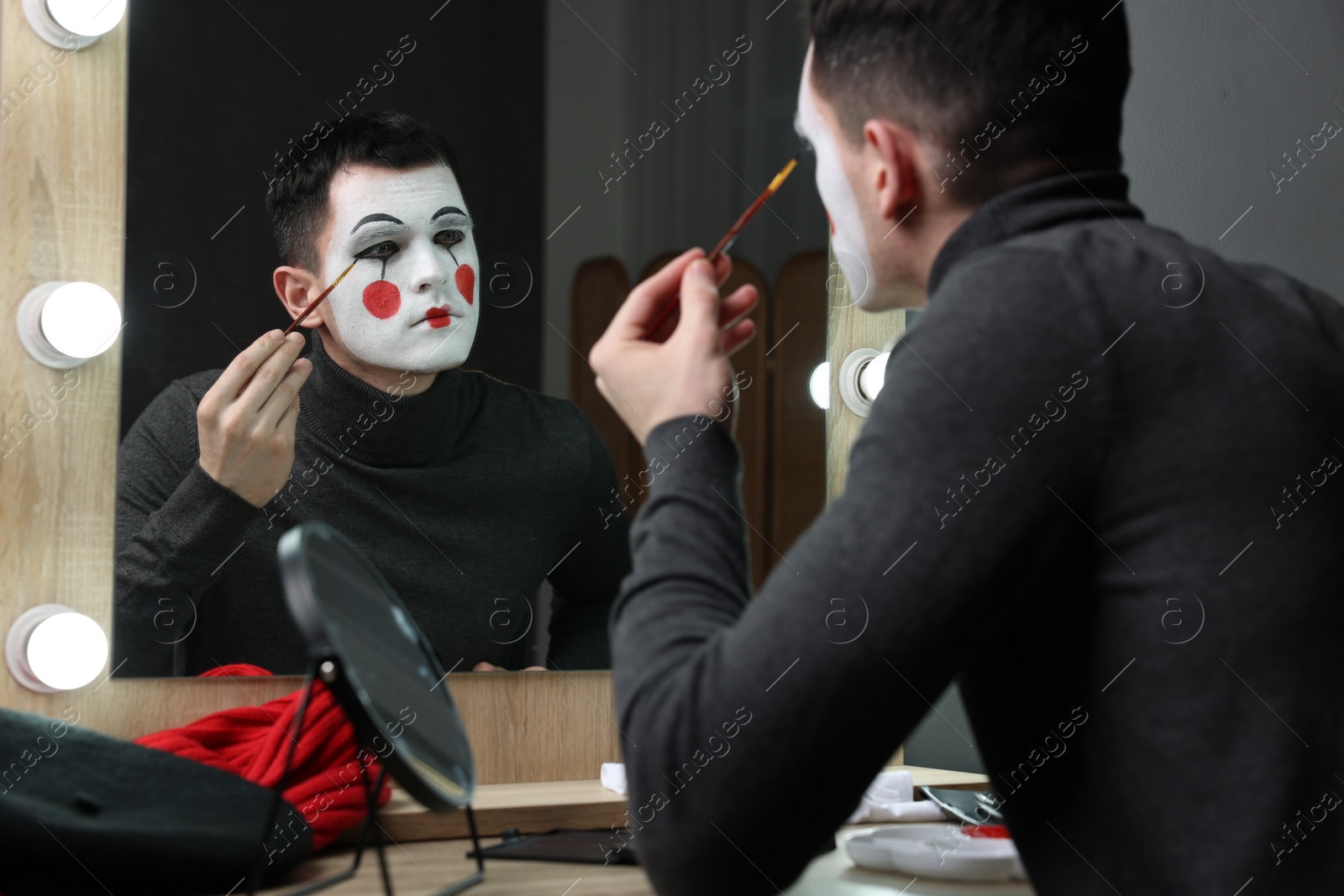 Photo of Young man applying mime makeup near mirror in dressing room