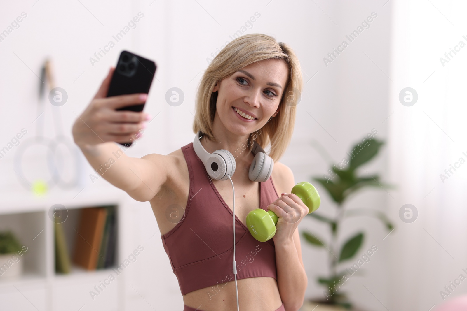 Photo of Smiling sports blogger holding dumbbell while streaming online fitness lesson with smartphone at home