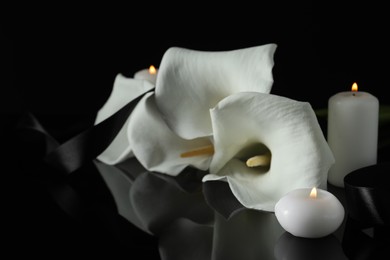 Photo of Burning candles, ribbon and white calla lily flowers on black mirror surface in darkness, closeup. Funeral symbols