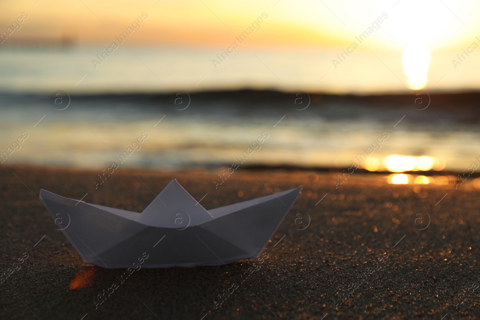 Photo of White paper boat on sand near sea at sunset, closeup. Space for text