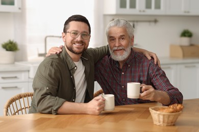 Photo of Happy son and his dad with cups at wooden table in kitchen