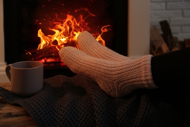 Photo of Woman in warm socks with cup of hot drink resting near fireplace at home, closeup
