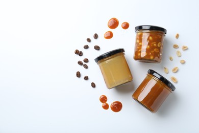 Photo of Tasty salted caramel in jars with peanuts and coffee beans on white background, flat lay. Space for text