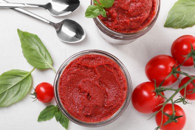 Photo of Jars of tasty tomato paste, spoons and ingredients on white table, flat lay