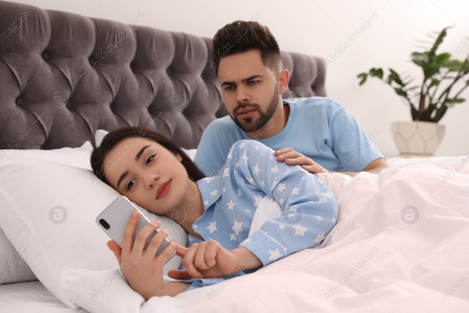 Photo of Young woman ignoring her distrustful boyfriend while using smartphone in bed at home