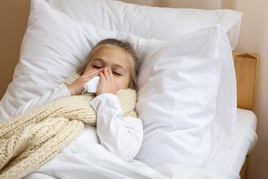 Photo of Sick girl with scarf and tissue lying in bed while blowing nose indoors