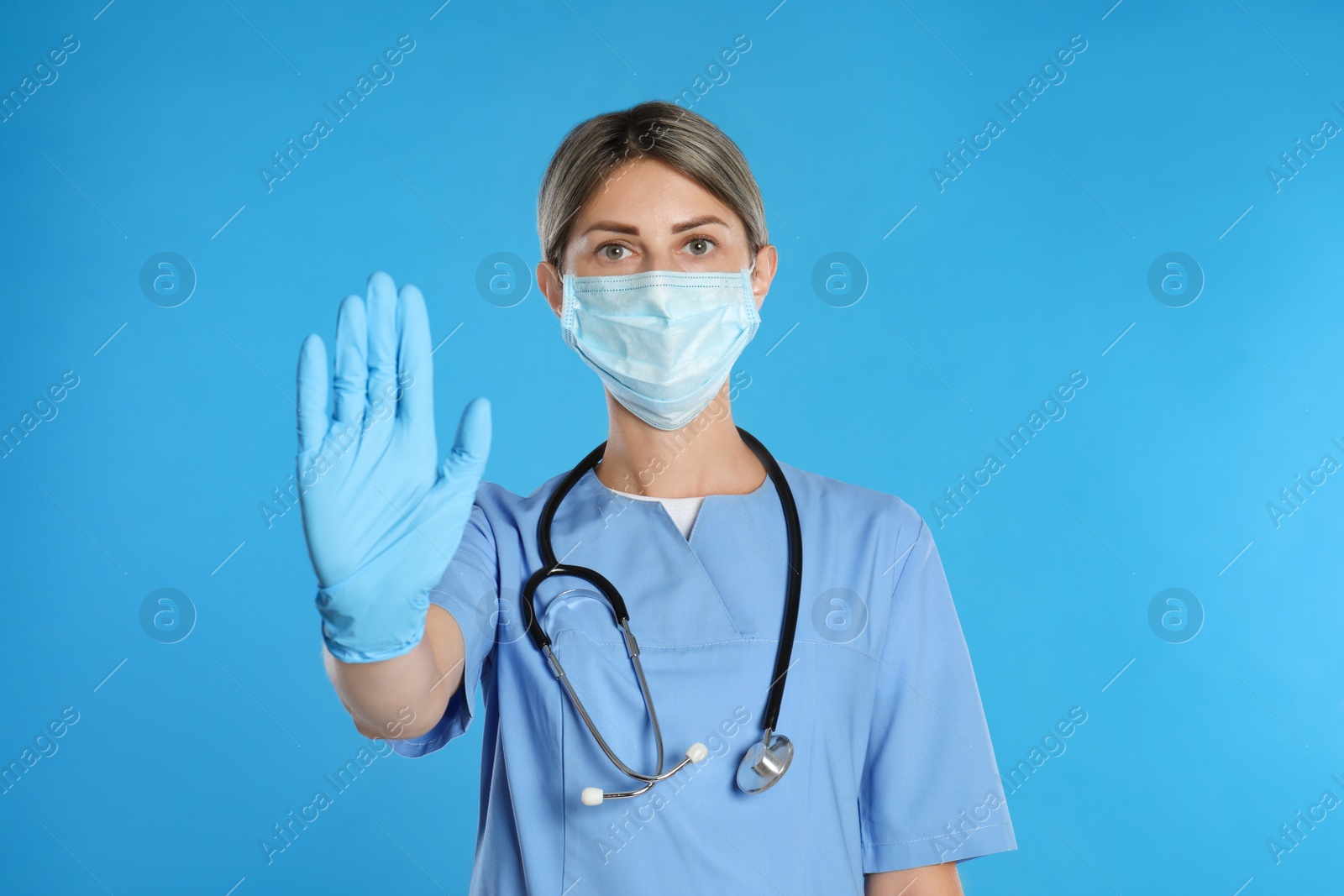 Photo of Doctor in protective mask showing stop gesture on light blue background. Prevent spreading of coronavirus