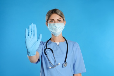 Photo of Doctor in protective mask showing stop gesture on light blue background. Prevent spreading of coronavirus