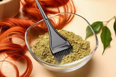 Photo of Bowl of henna powder, brush, green leaves and red strand on beige background, closeup. Natural hair coloring