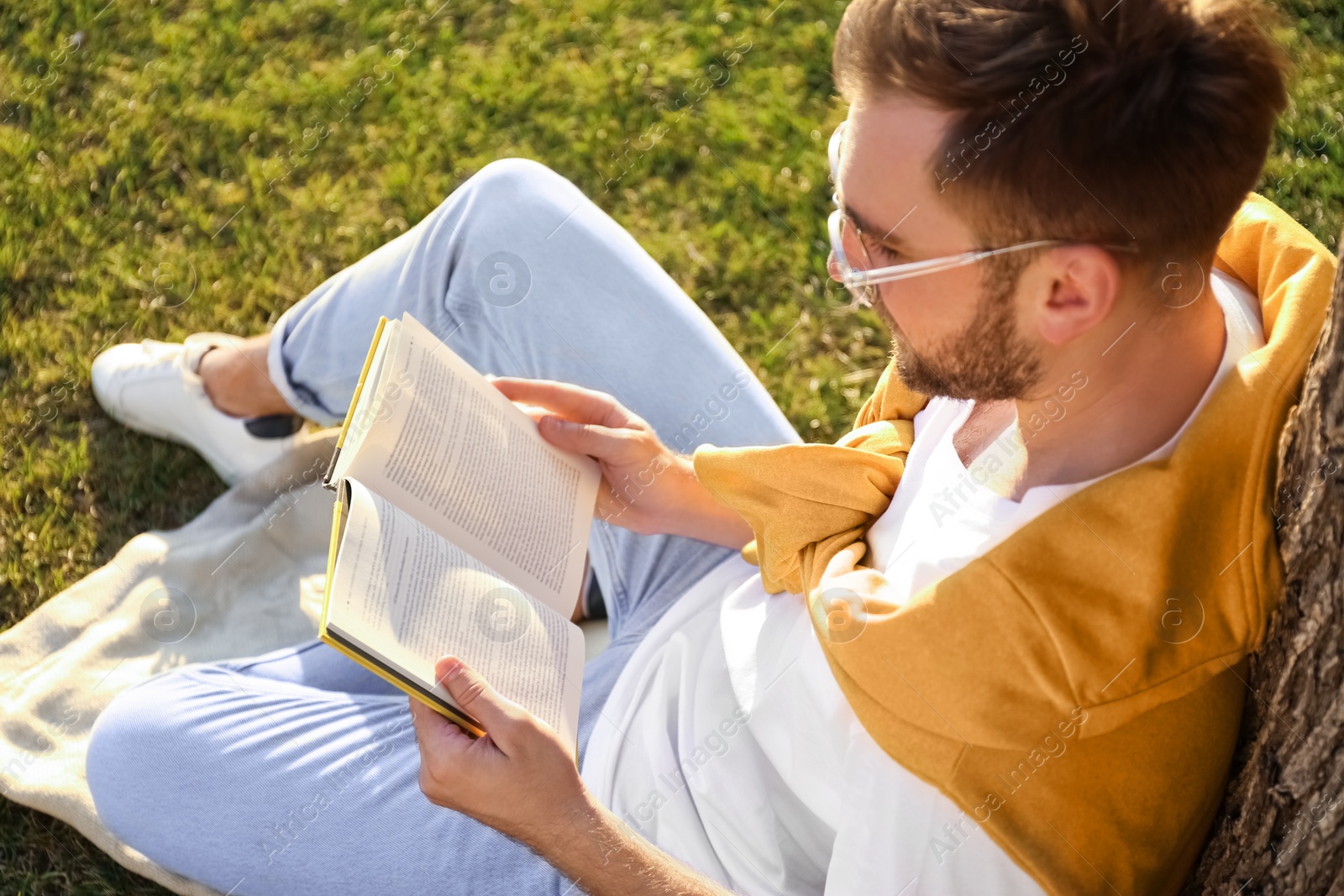 Photo of Young man reading book on green grass near tree in park