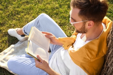 Young man reading book on green grass near tree in park