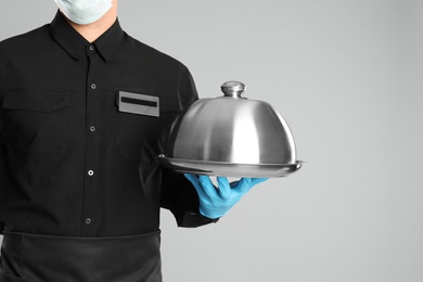 Photo of Waiter in medical face mask holding tray with lid on light grey background, closeup