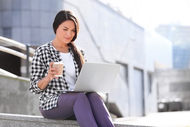 Beautiful woman with cup of coffee using laptop on city street