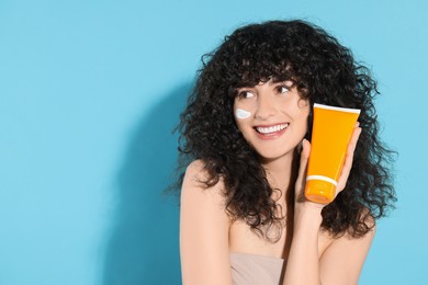 Photo of Beautiful young woman with sun protection cream on her face holding tube of sunscreen against light blue background, space for text