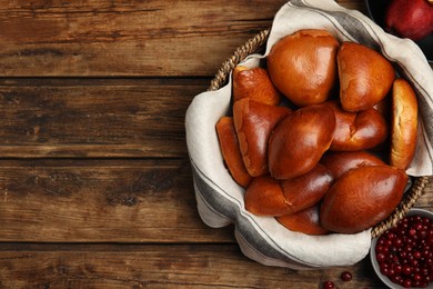 Photo of Delicious baked pirozhki in wicker basket, apple and cranberry on wooden table, flat lay. Space for text